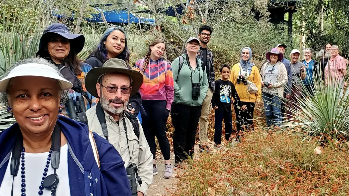 A group of smiling birders of all ages, many with binoculars, lined up along a garden trail