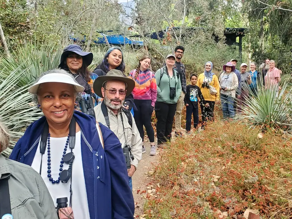 A line of smiling birders of all ages, many with binoculars, on a garden trail