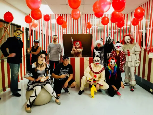 Colleagues dressed as clowns for Halloween, in front of red-and-white stripes and red balloons.